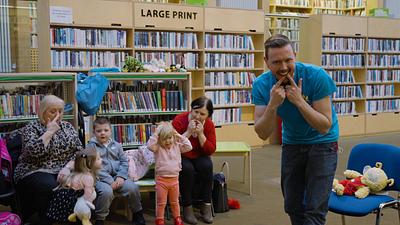 A man in a blue Bookbug t-shirt gesturing to library guests and encouraging them to smile